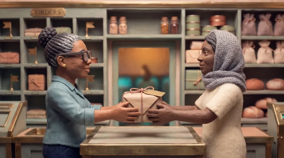 Nomsa and a customer either side of the shop counter, both smiling at each other, a small string-tied parcel passing between their four hands. The counter beneath is completely bare: no ledger, no paper, nothing being looked up. Behind them the shelves are stocked and through an open doorway the machines glow softly teal, running and unattended